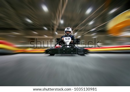 The man is going on the go-kart on karting track indoors. He is wearing a helmet.