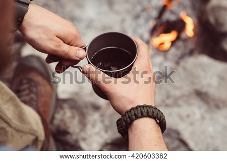 Man traveler hands holding cup of tea near the fire outdoors. Adventure, travel, tourism and camping concept. Hiker drinking tea from mug at camp. Coffee cooked over a campfire on the nature.