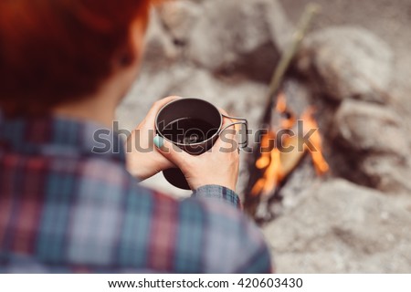 Woman traveler hands holding cup of tea near the fire outdoors. Adventure, travel, tourism and camping concept. Hiker drinking tea from mug at camp. Coffee cooked over a campfire on the nature.