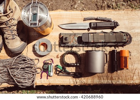 Overhead top view of hiking travel gear on wood log. Items include hiking boots, cup, rope, knife, matches, flashlight, compass. Flat lay of outdoor travel equipment items for mountain camping trip.
