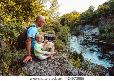 Father and son sitting near beautiful river on nature in summer. Family spending time together. Dad with kid, little baby boy travel outdoors. Man and child traveling in nature park, forest, river.