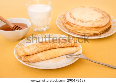 Pancakes rolled in a tube and poured honey on a plate, a glass of milk, a bowl of honey, a plate with a stack of pancakes. Pancakes and honey on the yellow tablecloth.\
Horizontal shot. Close-up.