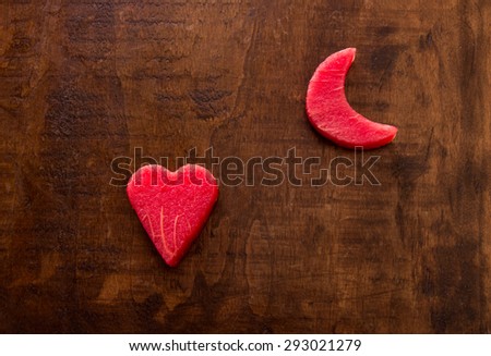 On the left side of the frame a heart of watermelon flesh in the upper right the moon on a wooden background. Watermelon heart and moon. Top view. Close-up. Daylight. Horizontal shot.