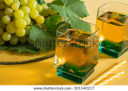On the left grapes with leaves on a plate,  and on the right a 2 glasses of grape juice on the yellow background. 2 glasses of grape juice. Horizontal shot. Daylight.