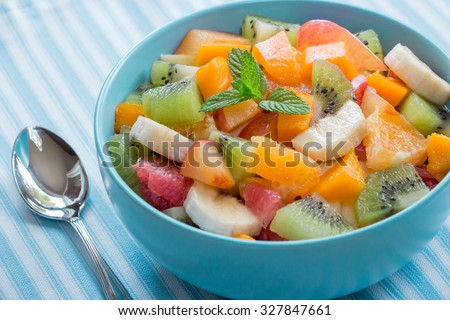 In the frame blue plate with fruit salad of oranges, bananas, grapefruit, plum, kiwi, mango on a background of blue cloth with white stripes. Fruit salad. Daylight. Horizontal shot.