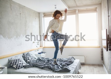young happy woman jumping on bed in morning, stylish bedroom, positive mood, wearing jeans and knitted white sweater, cheerful, active life, energy, having fun, slim body, smiling, happy
