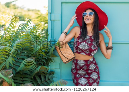stylish beautiful woman in red hat posing on blue background, printed outfit, summer style, fashion trend, top, skirt, skinny, straw handbag, sunglasses, accessories, smiling, happy, tropical vacation