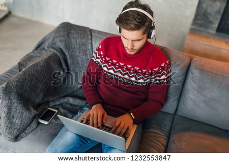 young attractive serious concentrated man sitting on sofa at home in winter, wearing red knitted sweater, working on laptop, freelancer, busy, listening to headphones, typing, studying online