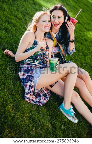 two young beautiful happy stylish hipster girls,  friends together, cocktail, drink, denim outfit, smiling, happy, fashion, cool accessories, vintage style, having fun, park, sitting, grass, laughing