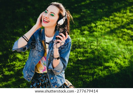 young hipster stylish beautiful girl listening to music, mobile phone, headphones, enjoying,  denim outfit, smiling, happy, cool accessories, vintage style, having fun, laughing, park