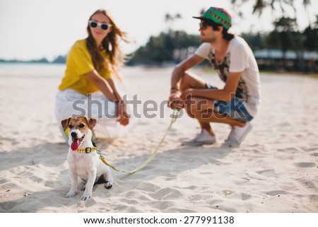 young stylish hipster couple in love walking playing dog puppy jack russell in tropical beach, white sand, cool outfit, romantic mood, having fun, sunny, man woman together, horizontal, vacation