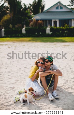 young stylish hipster couple in love walking playing dog puppy jack russell in tropical beach, cool outfit, romantic mood, having fun, sunny, man woman together, vacation, house home villa background
