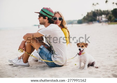 young stylish hipster couple in love walking playing dog puppy jack russell in tropical beach, white sand, cool outfit, romantic mood, having fun, sunny, man woman together? horizontal, vacation