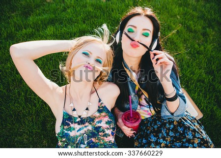 two young beautiful stylish hipster girls, friends together, cocktail, drink, denim outfit, smiling, fashion, cool accessories, vintage style, having fun, park, crazy, hair, mustache, listening music