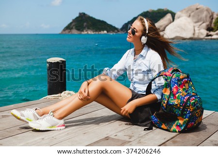 young stylish hipster woman traveling around the world, sitting on the pier, aviator sunglasses, headphones, listening to music, vacation, backpack, denim shirt, happy, tropical island lagoon