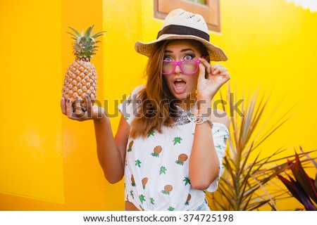 young beautiful stylish hipster woman, straw hat holding pineapple, pink sunglasses,  funny, happy summer, yellow wall background, denim shorts, cool t-shirt, trendy outfit, tropic, shocked, surprised
