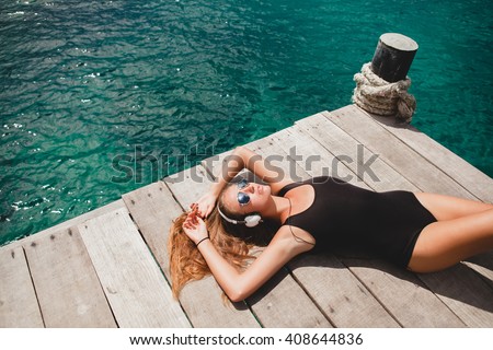 young slim woman laying on pier, Mediterranean Sea, azure water, sunny, tanned skin, listening music, headphones, black swimsuit, sexy body, sunbathing, tropical vacation, relaxed, sunglasses
