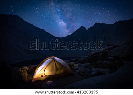 An illuminated tent in the outdoors with the milky way and stars rising in the background.
