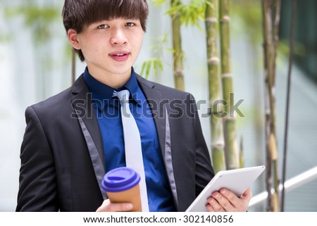Young Asian male business executive using tablet PC and holding coffee cup smiling