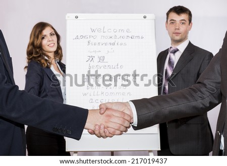 Male handshake on the foreground, male and female corporate trainers on background. Flip chart with WELCOME sign written in many different languages. Focus on hands, but flip chart signs are visible.