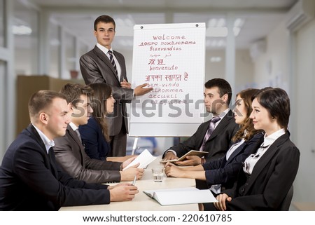 Young corporate coach facilitates the brainstorming of the team of six people. He use flip chart, attendants are sitting around the table and discussing the task
