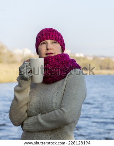 Warmly dressed European lady is standing in front of the sea or river bay and keeping white coffee mug