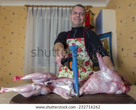 Smiling cook carves a rabbit at home kitchen. Male cook dressed in colorful apron cuts rabbit by axe at regular home kitchen