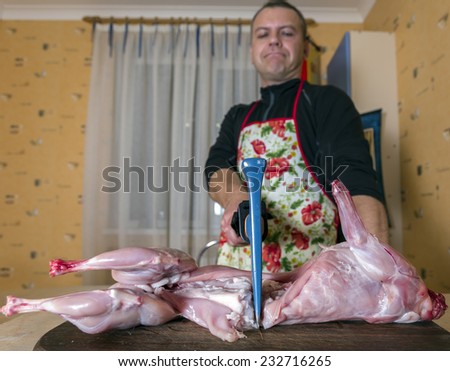 Serious cook carves a rabbit at home kitchen. Male cook dressed in colourful apron cuts rabbit by axe at regular home kitchen