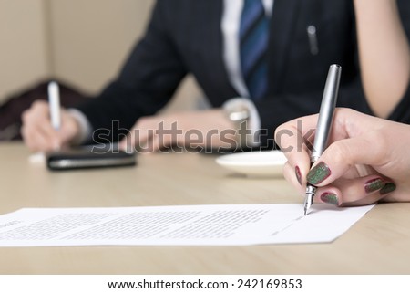 Female signs contract. Close up of female hand signing formal paper on the office table. The business counterpart on the background