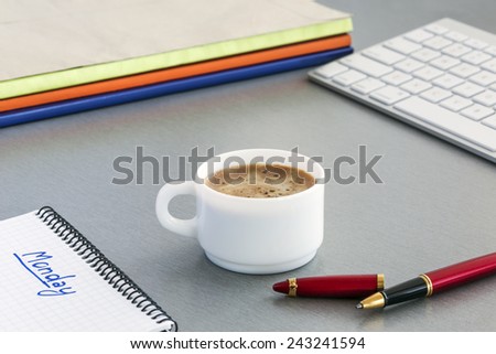 Office working place. Workspace on grey wooden table surrounded with red pen, computer keyboard, booklets and notepad with sign MONDAY. Coffee cup in the middle. Focus is on the coffee surface
