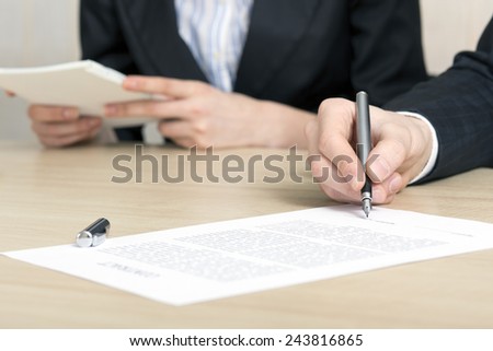 Male businessperson signs contract. Close up of female hand signing formal paper on the office table. The business counterpart on the background.