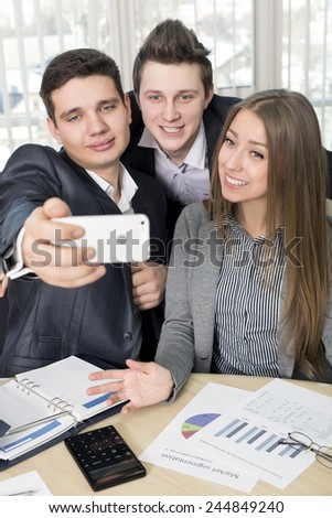 Happy co-workers makes selfie. Three laughing young business people makes SELFIE with the sell phone. Office interior, stationery, paper, charts on the table