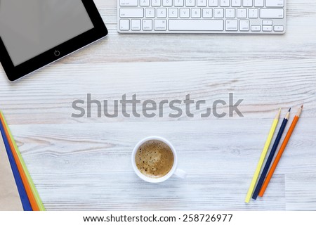 Creative working desk with coffee mug in the center. \
Top view on working place with cup of coffee, tablet PC, computer keyboard, and color matching booklets and pencils.
