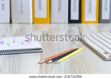 Side view on the working place of designer.\
Light wooden desk with color pencils, notepad with marked handwriting, computer keyboard and line of folders on the background.