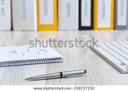 Side view on the working place of manager.\
Light wooden desk with color pen, notepad with marked handwriting, computer keyboard and line of folders on the background