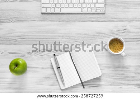 Business break.\
Composition with notepad, keyboard, coffee mug with thick foam, green apple, located on wooden desk. Everything is black and white except for apple and coffee.