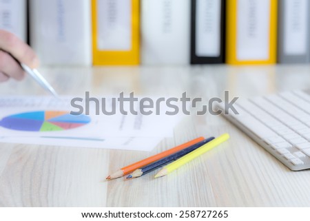 Manager working on presentation.\
Side view of desk with keyboard, pencils, papers with charts and human hand keeping pen. Stack of multicolored office folders on the background.