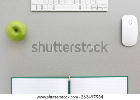 Working space in grey-green. Creative office style composition with green apple, computer keyboard and mouse, big opened note pad with green pencil, located on grey wooden desk. Top view.