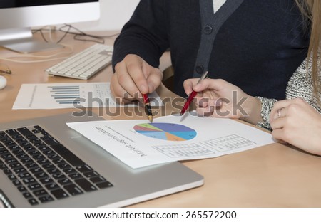 Hands of people working on printed charts.\
Palms of male and female pointing with pens on the printed colorful charts. Office desk with laptop keyboard on the foreground and large screen on background