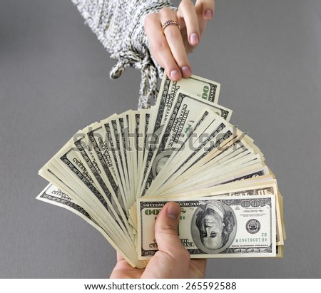Taking cash note of the hands of husband.\
Female hand pulls out single cash note from the large stack of notes held by male hand. Caucasian people, grey wooden desk on the background