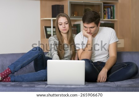Home entertainments.\
Young Caucasian man and woman watch movie on the laptop. Home interior, blue sofa, casual dress code