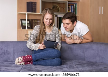Home entertainments.\
Young Caucasian man and woman watch movie on the tablet PC. Home interior, blue sofa, casual dress code