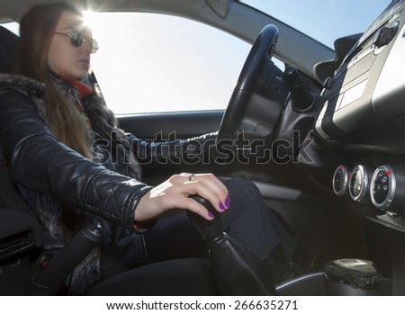 Confident female driver.\
Young woman drives car, focus on her hand switching transmission grip. Steering wheel on the right, blues sky in the window, bright sunbeams on the upper.