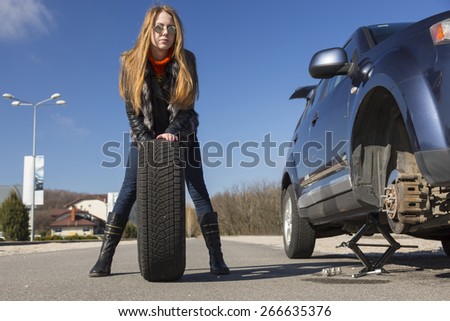Female driver repairs car.\
Young female, dressed biker style, rolls big wheel towards broken car.
