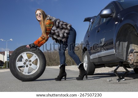 Female driver repairs car.\
Young female, dressed in jeans pants, red sweater and fur vest, rolls big wheel from the broken car.