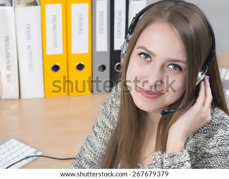Young smiling customer support operator. Portrait of smiling beautiful lady with headset on. Smart casual dress, stack of colorful office folders on the background.