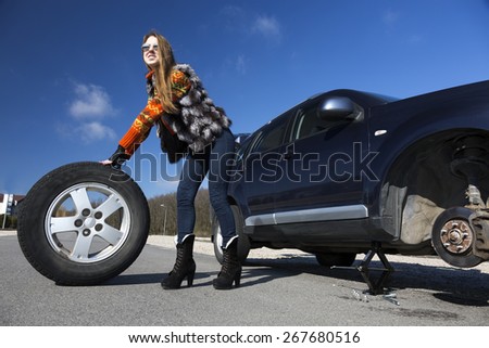 Female driver repairs car. Young female, dressed in jeans pants, red sweater and fur vest, rolls big wheel away from the broken car. Bright blue sky, sun shining day