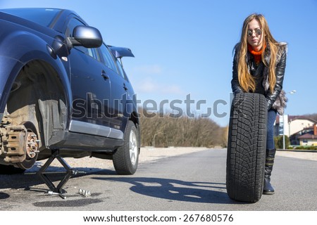 Female driver repairs car. Young female, dressed in jeans pants, black leather jacket and fur vest, rolls big wheel towards  broken car