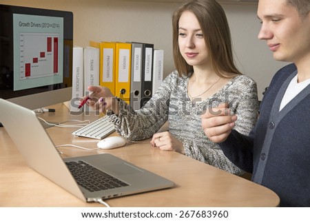 Exchange of opinions. Office workers discuss the task sitting at the desk and gesturing. Smiling, friendly faces. Opened laptop on the foreground, large screen on the background
