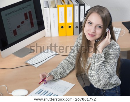 Young businesswoman with headset on in the office interior. Portrait of smiling cute lady with headset sitting at the office desk. Colored printed charts, folders and desktop are located on the desk